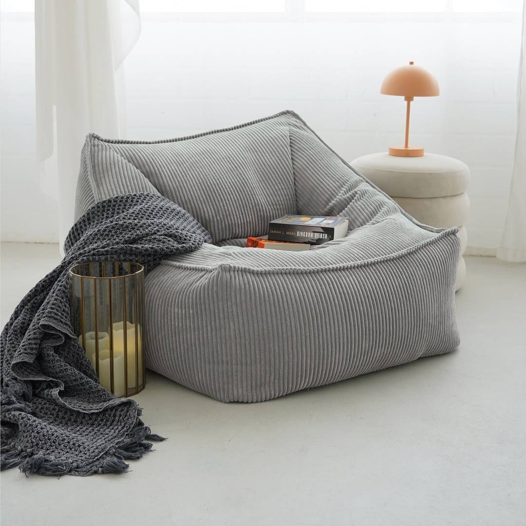 Pouf-lit Bean Bag en velours côtelé gris clair 'Der Lümmel' dans le salon avec une couverture, deux livres posés sur le pouf.
