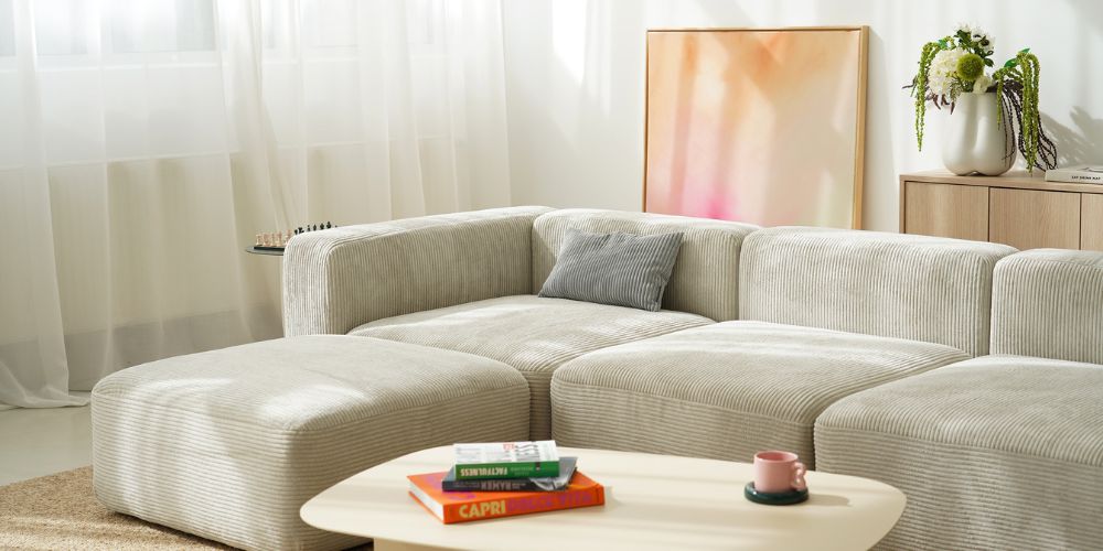 Canapé modulaire en velours côtelé beige en forme de L avec coussin gris, devant une table basse ovale blanche avec des livres et une tasse rose, rideaux clairs en arrière-plan et tableau abstrait rose-orange.