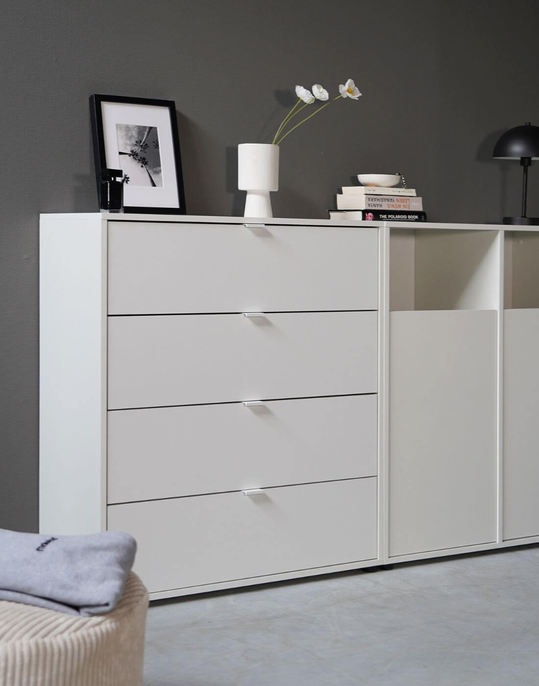 Commode blanche 'Die Stilvolle' avec quatre tiroirs à côté d'une armoire à linge blanche 'Der Wäschehüter' contre un mur gris