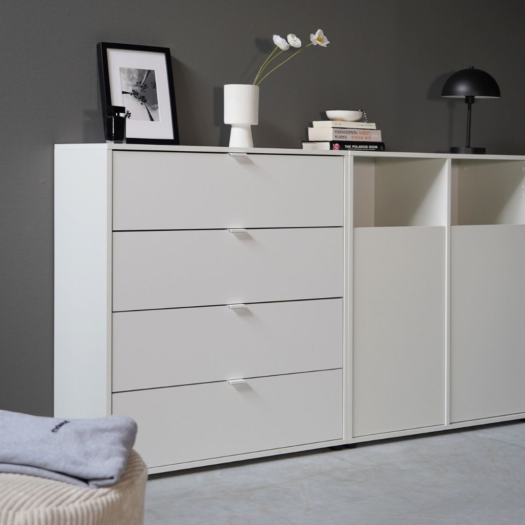 commode blanche 'Die Stilvolle' avec quatre tiroirs à côté d'une armoire à linge 'Der Wäschehüter' avec deux portes contre un mur gris